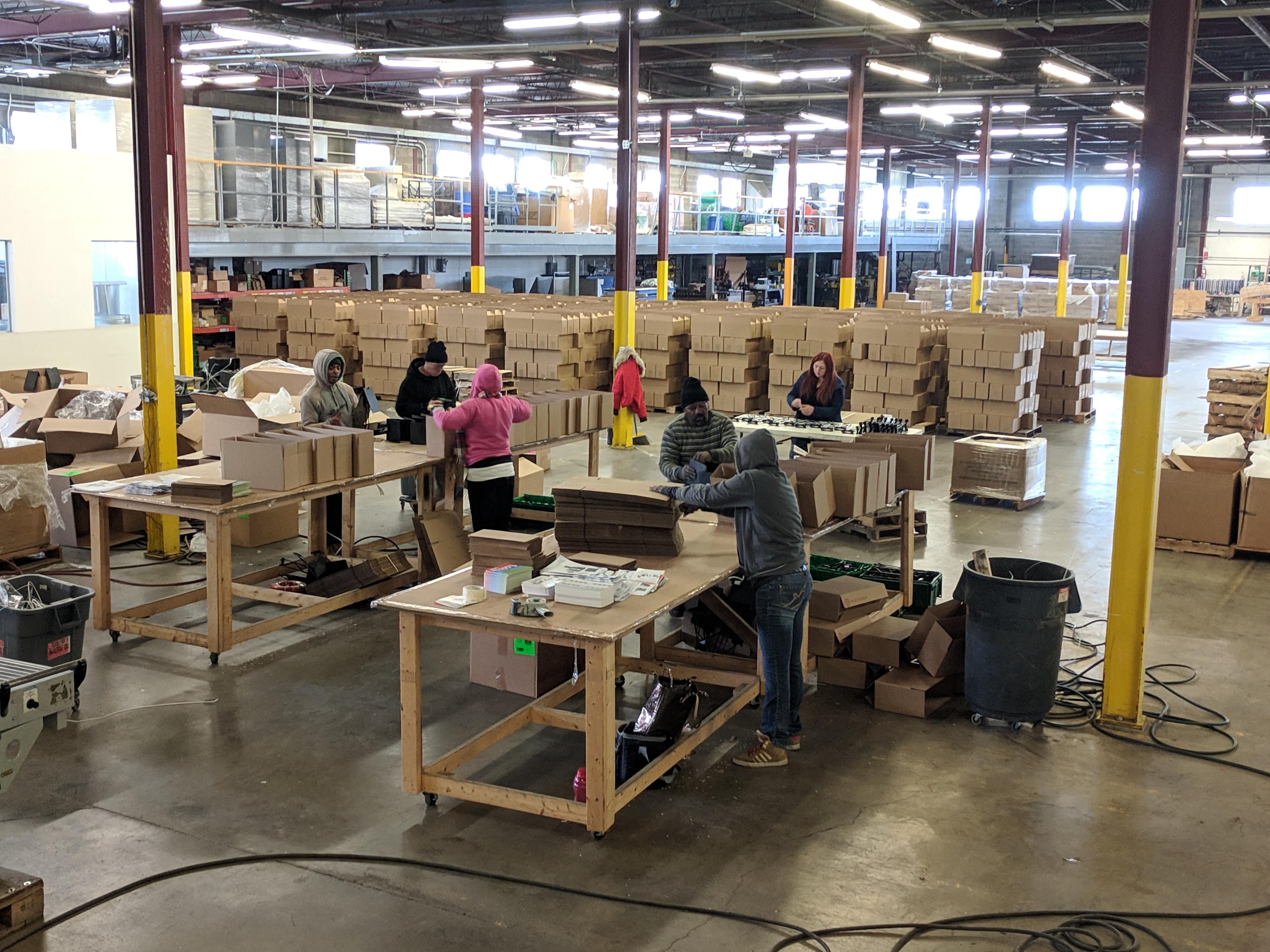 People on an assembly line with pallets of boxes behind them inside a factory